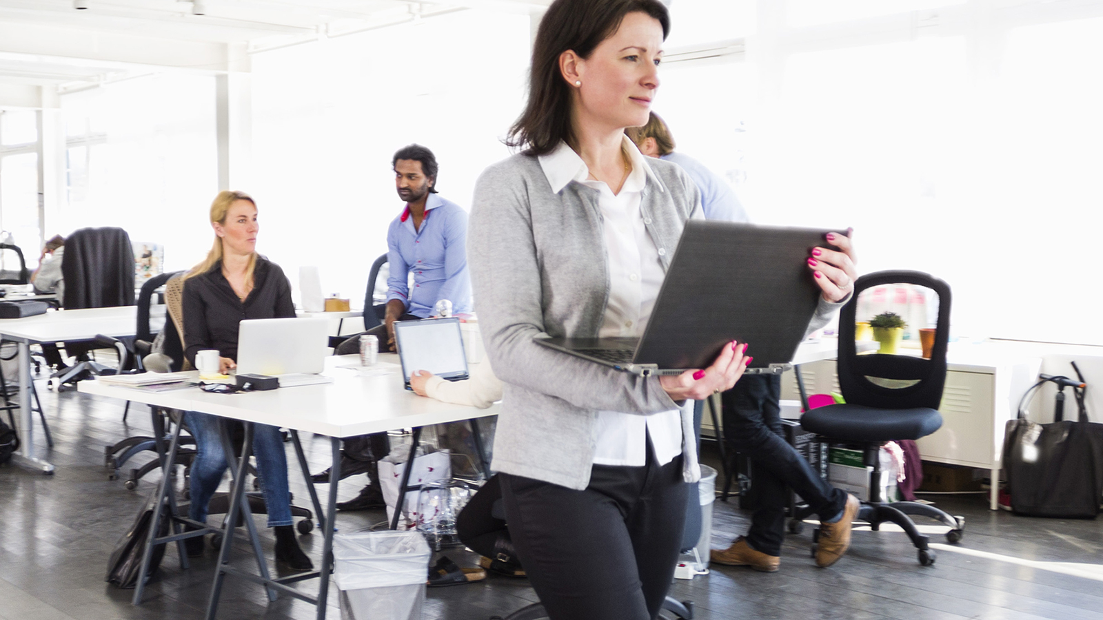 Woman with laptop walking among office tables.