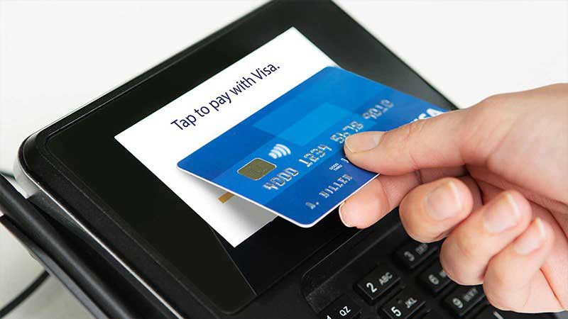 A Visa credit card is being held by a hand, ready to pay contactless at a POS terminal. 