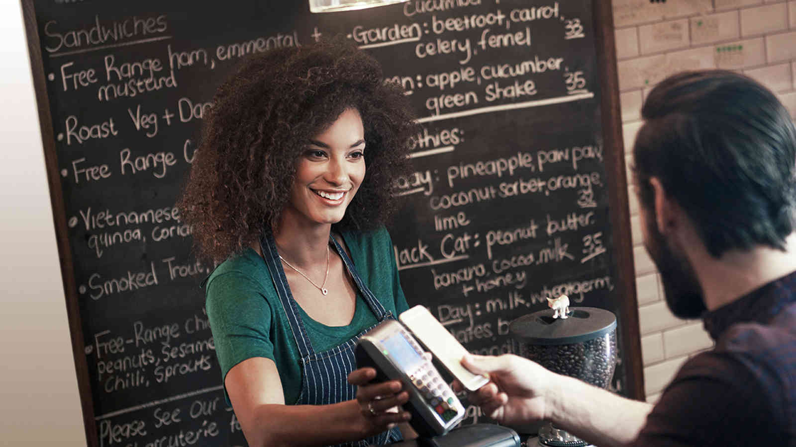 mobile-checkout-1600x900 Woman holding a payment terminal for a man who is making a contactless mobile payment.