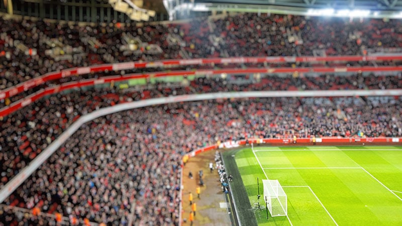 Blurred background of football players playing and soccer fans in match day on beautiful green field with sport light at the stadium