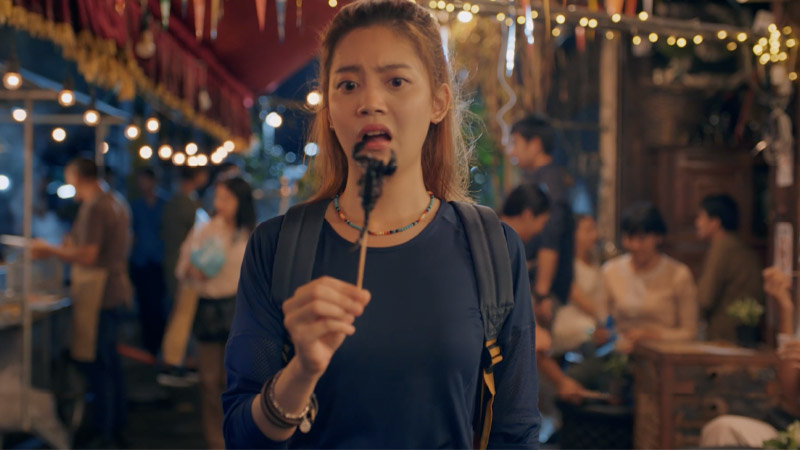 A young lady on a street food market holding a stick with a scorpion on it, which she bought with her travel payment card.