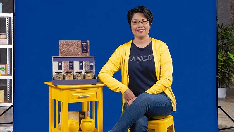 A woman sits on a stool next to a table with rice promoting the help for small businesses. 