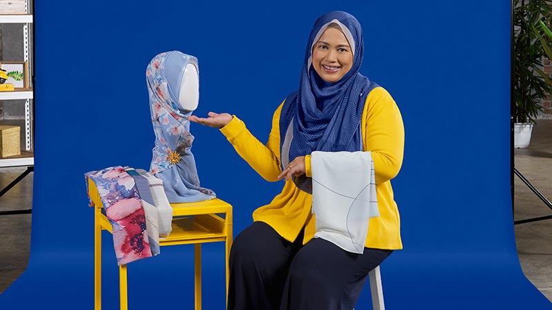 A woman wearing a yellow shirt and blue hijab sits on a chair, promoting help for small businesses. 